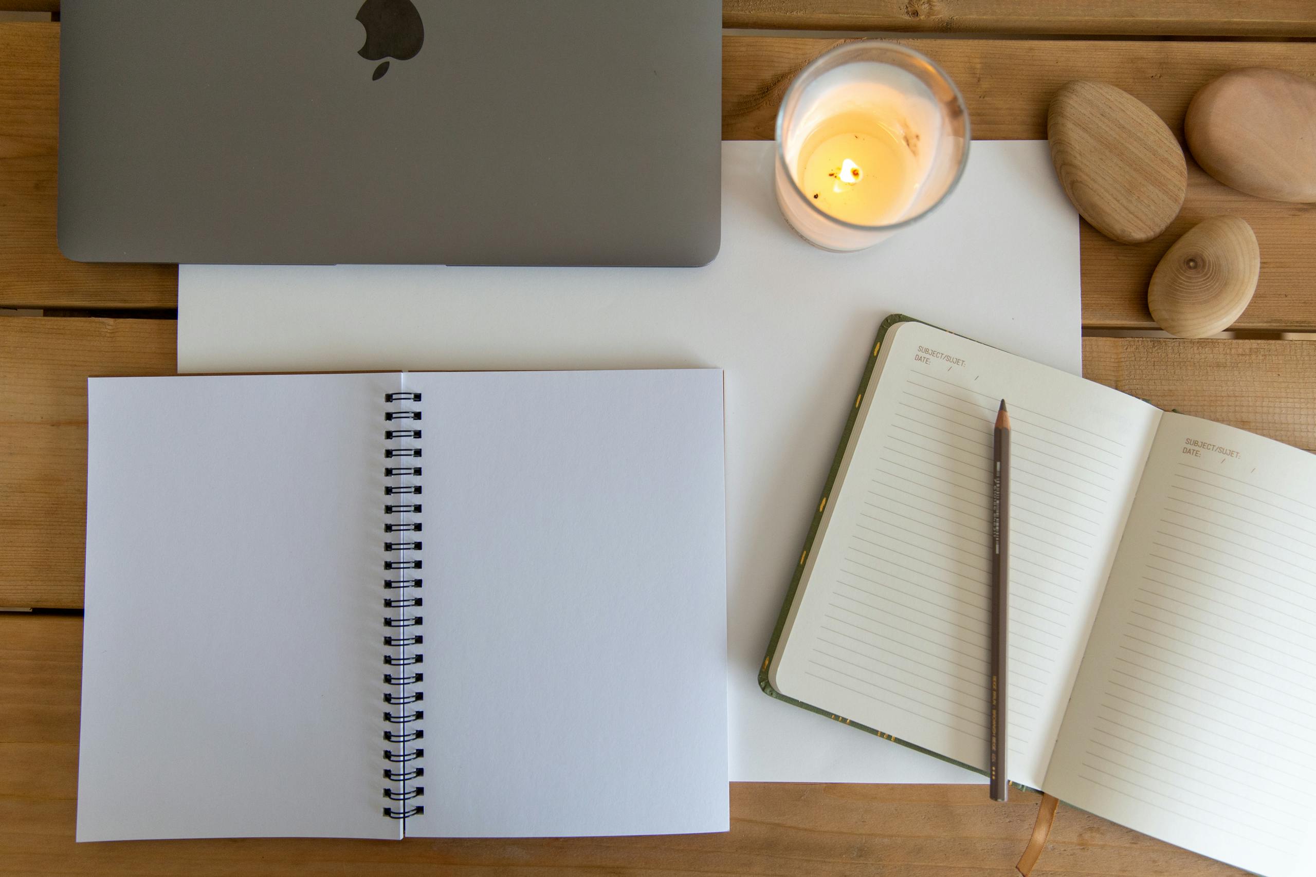 A serene workspace featuring a laptop, open notebooks, and a lit candle on a wooden surface.